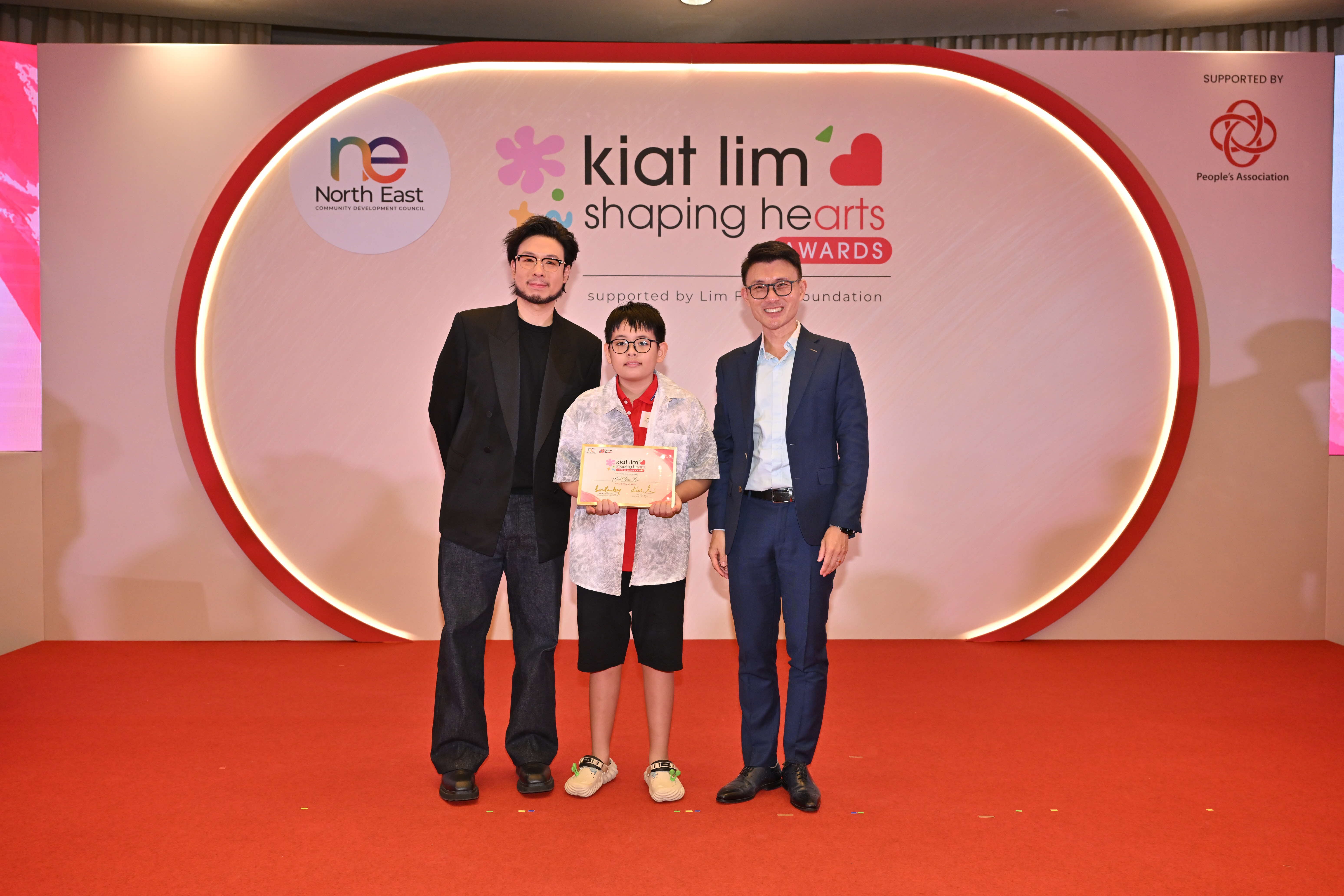 Award recipient standing on stage with the presenter, holding their certificate and posing for a group photo during the award ceremony, with a backdrop reading ‘Kiat Lim-Shaping Hearts Awards'.